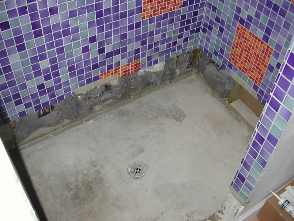 Douche en cours de rénovation après dépose du carrelage de sol, chape à nu et murs couverts de mosaïque violette et orange.
